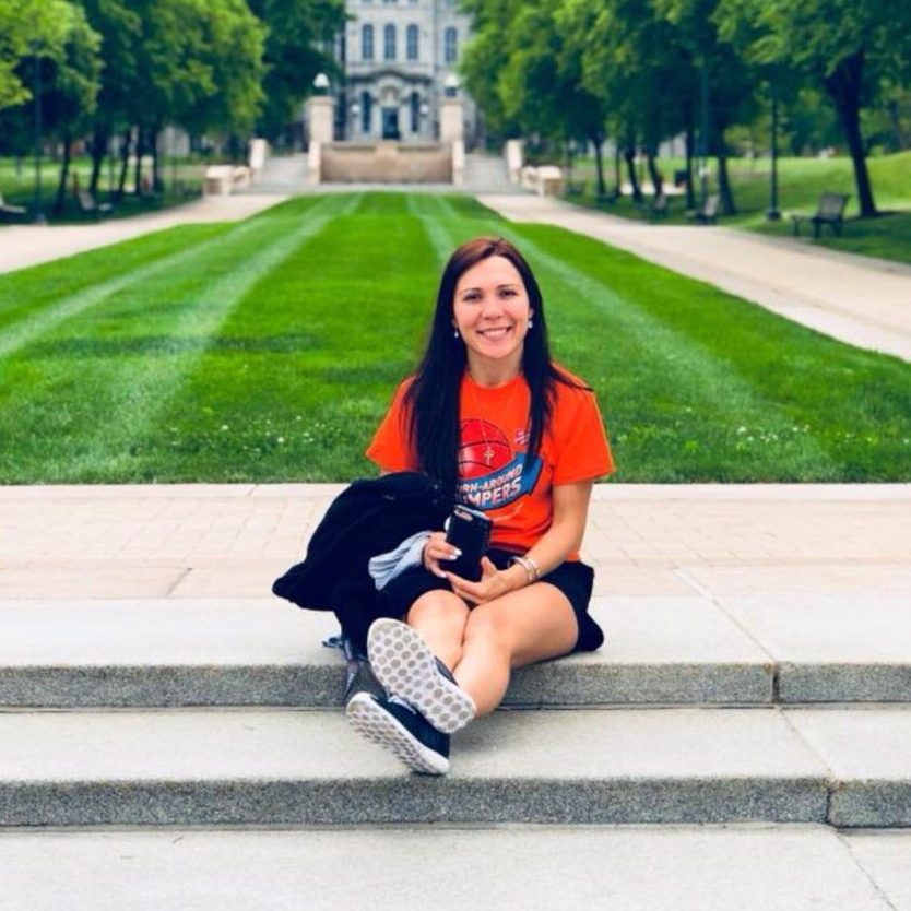 A young woman sitting on stone steps wearing an orange t-shirt and black shorts, smiling at the camera. In the background, there is a well-manicured lawn and trees, with a historic building visible at the top of the stairs.